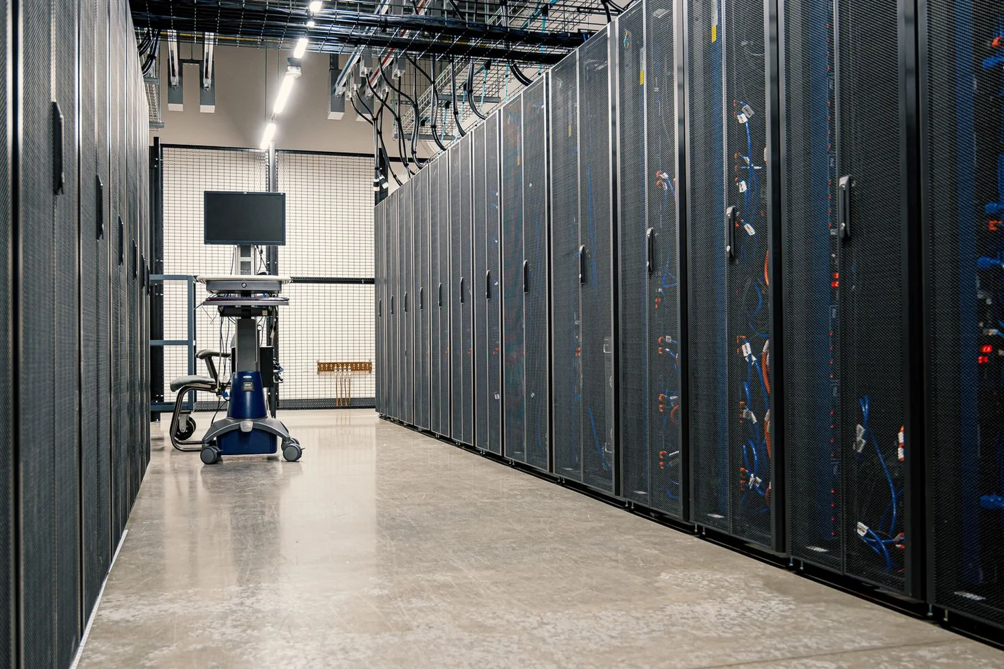 Rows of enclosed server racks beneath overhead cable trays in a data centre aisle